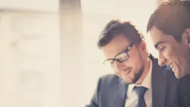 Two people in suits smiling and working together, sitting near a window.