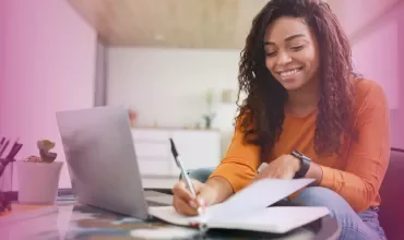 Person smiling while taking notes at a desk with a laptop.